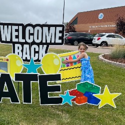 A kindergartner stands next to a Welcome Back sign in front of our school.